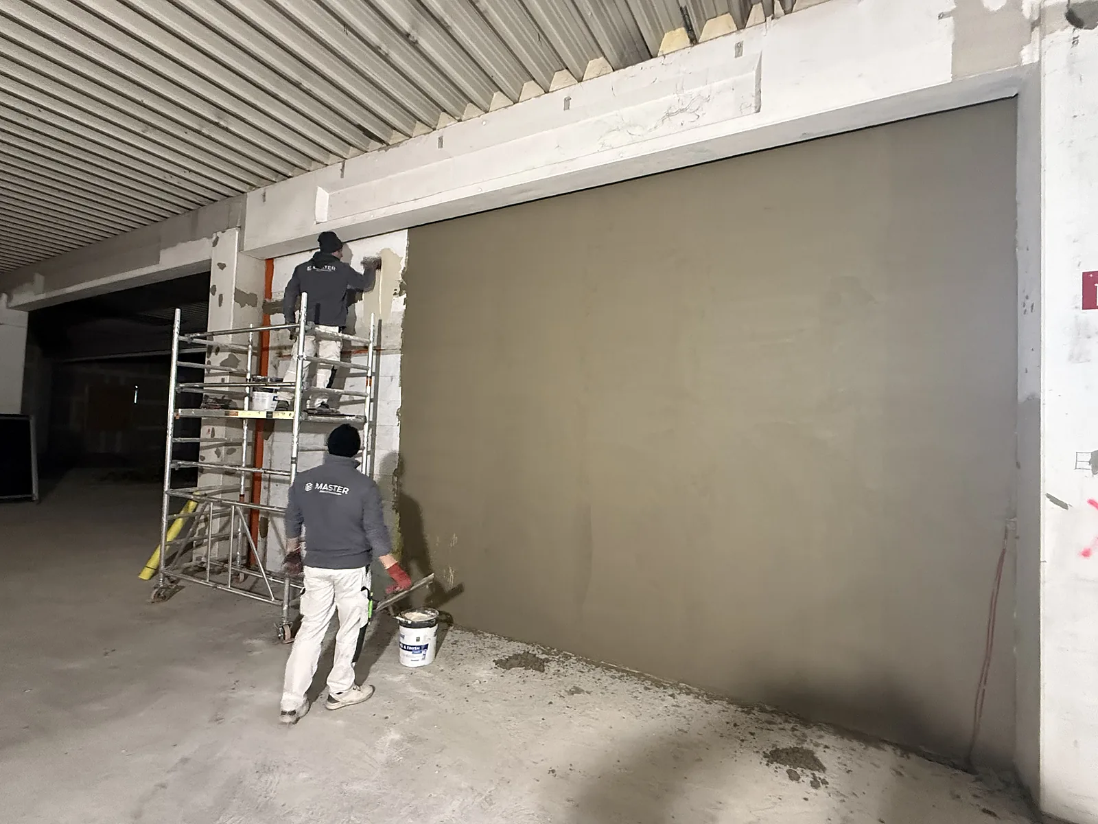 Zwei Handwerker in MASTER-Arbeitskleidung arbeiten an einer großen Wand, einer davon auf einem Rollgerüst